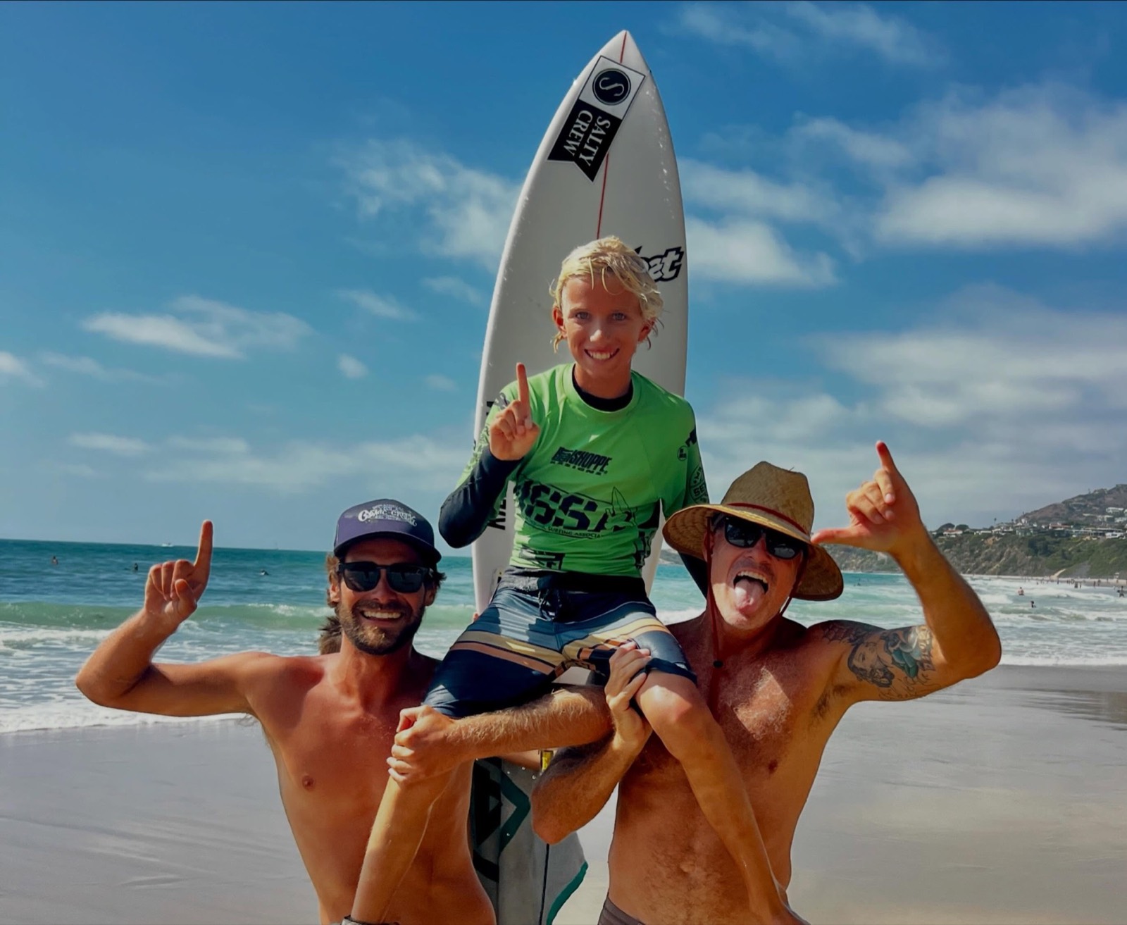 Two coaches carrying a smiling young surfer on their shoulders at the beach